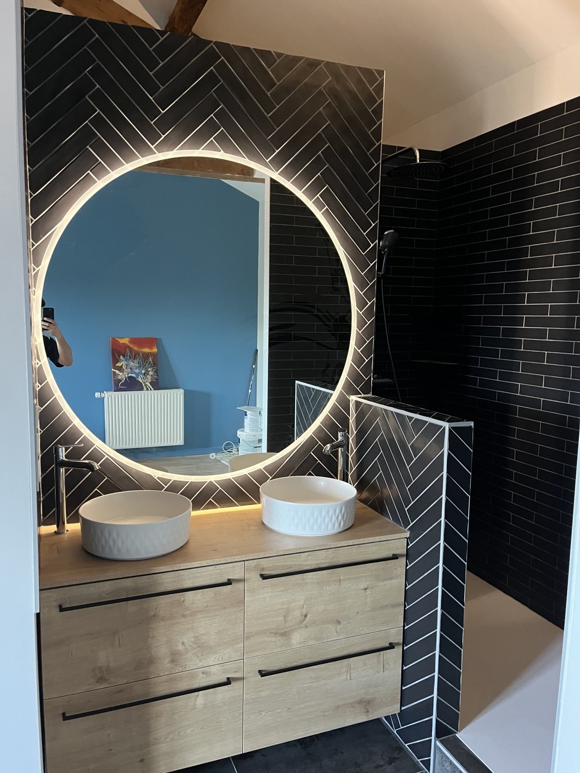 Salle de bain avec carrelage noir en chevrons, double vasque blanche, meuble en bois et grand miroir rond lumineux