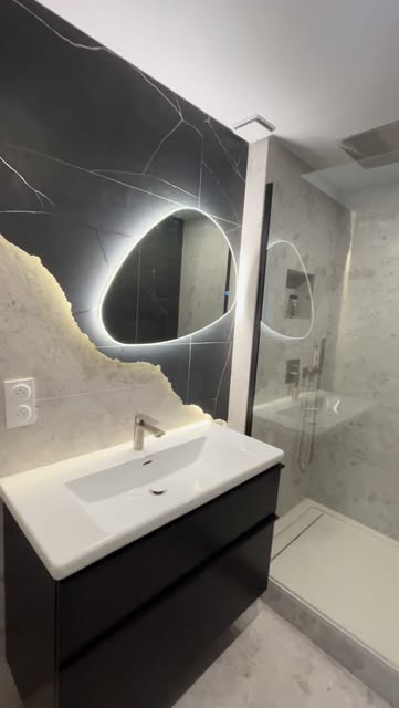 Salle de bain rénovée, vasque blanche, mur sombre et miroir rond lumineux